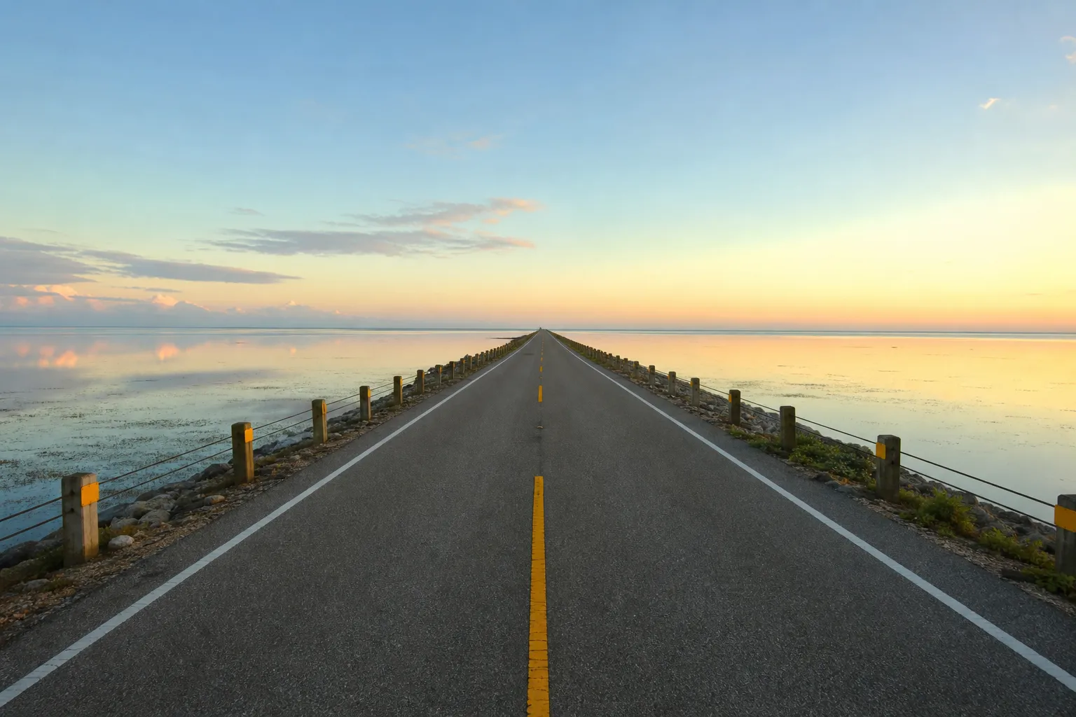 A straight, empty coastal causeway road running toward a bright horizon with shallow Gulf water on both sides in soft morning light.