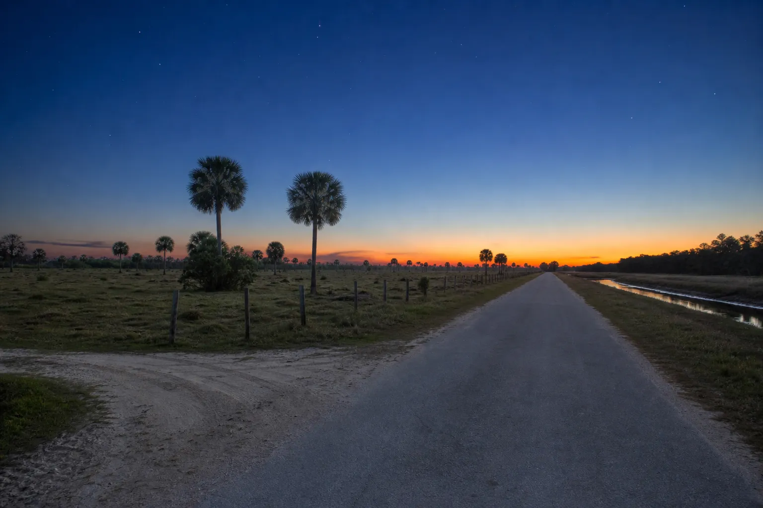 A straight rural Florida county road at dusk between cattle pasture and a canal, with a sandy side track joining the pavement.
